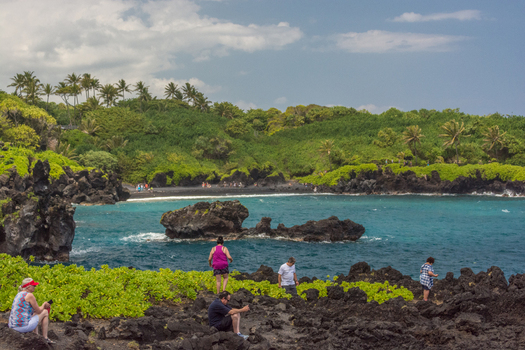 View of the black sand beach at Waianapanapa State Park with volcanic rock formations turquoise water and lush tropical vegetation in Maui County Hawaii United States.