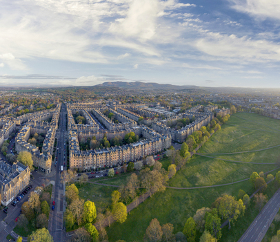 Aerial view of historic stone terraced houses bordering a lush green park under a soft blue sky with distant hills Edinburgh Scotland United Kingdom. Aerial view of historic stone terraced houses bordering a lush green park under a soft blue sky with distant hills Edinburgh Scotland United Kingdom.