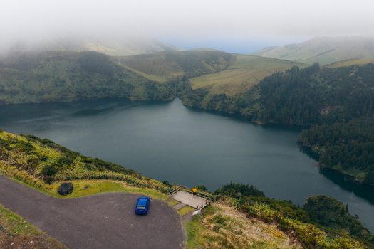 Aerial view of Caldeira Funda volcanic lake surrounded by lush green hills and mist with a blue car parked at a viewpoint in Lajes das Flores Azores Portugal. Aerial view of Caldeira Funda volcanic lake surrounded by lush green hills and mist with a blue car parked at a viewpoint in Lajes das Flores Azores Portugal.