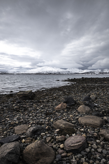 View of a rocky shoreline along the fjord with snow capped mountains under a cloudy sky in Tromso Troms Norway.