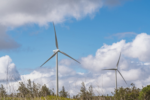 View of wind turbines at Parque Eolico da Serra dos Candeeiros set against a bright blue sky with scattered white clouds and green trees in the foreground in Rio Maior Santarem Portugal. View of wind turbines at Parque Eolico da Serra dos Candeeiros set against a bright blue sky with scattered white clouds and green trees in the foreground in Rio Maior Santarem Portugal.