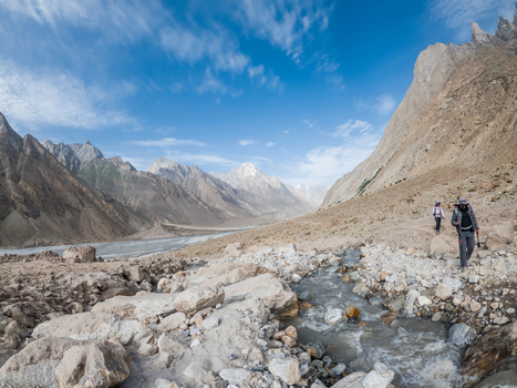 View of hikers trekking along a rocky path near a stream with the Baltoro Glacier and rugged mountain peaks in the background in Baltoro Gilgit Baltistan Pakistan.