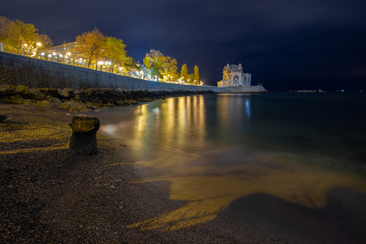 View of the Casino of Constanta illuminated at night along the Black Sea with reflections on the water and a dark pebbled beach in the foreground Constanta Judetul Constanta Romania. View of the Casino of Constanta illuminated at night along the Black Sea with reflections on the water and a dark pebbled beach in the foreground Constanta Judetul Constanta Romania.