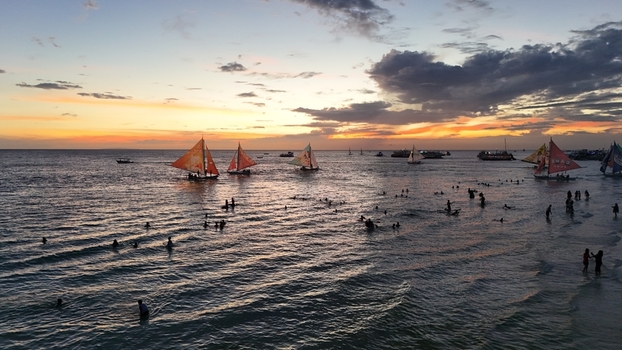 Malay Philippines 02 May 2026 Aerial view of Boracay Island with people swimming in the sea and traditional paraw sailboats under a vibrant sunset sky.