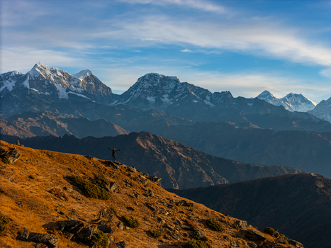 Aerial view of a person standing on a rocky ridge with arms outstretched against the backdrop of the snow capped Himalayan mountain range under a clear blue sky in Nepal.