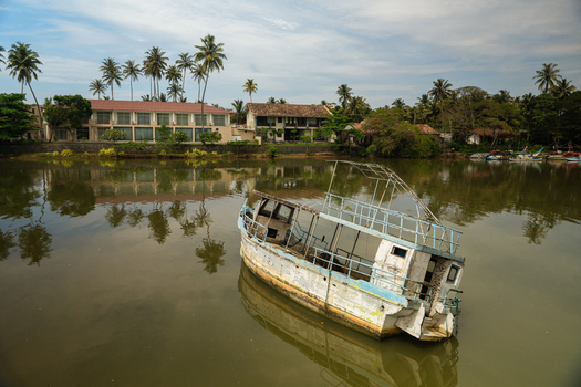 View of an abandoned wooden boat floating in a calm river surrounded by palm trees and coastal buildings in Mirissa Southern Province Sri Lanka. View of an abandoned wooden boat floating in a calm river surrounded by palm trees and coastal buildings in Mirissa Southern Province Sri Lanka.