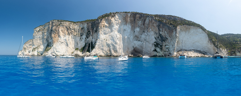View of sailboats anchored in the turquoise Ionian Sea in front of towering white limestone cliffs on the coast of Lefkada Greece.