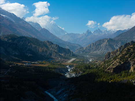 Aerial view of a mountain valley featuring a small airport runway a winding river dense pine forests and towering snow capped Himalayan peaks Nepal.
