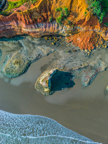 Aerial view of the rugged coastline with dramatic orange cliffs dark sand beach and gentle waves washing onto the shore in Bali Bali Indonesia. Aerial view of the rugged coastline with dramatic orange cliffs dark sand beach and gentle waves washing onto the shore in Bali Bali Indonesia.