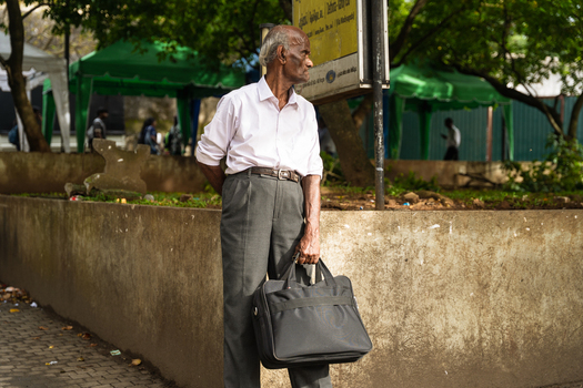 Sri Lanka 20 February 2026 View of an elderly man holding a black briefcase while standing near a concrete wall with green tents and trees in the background.