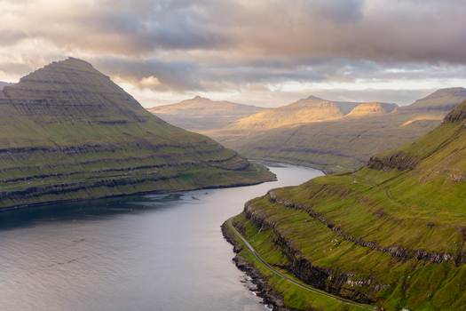 View of dramatic green mountains and a winding road along the fjord in the Faroe Islands. View of dramatic green mountains and a winding road along the fjord in the Faroe Islands.