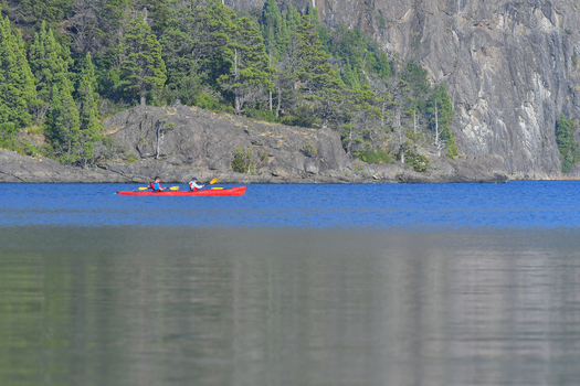 View of two people paddling a red kayak on the calm blue waters of Nahuel Huapi Lake with rocky shores and green trees SAN CARLOS DE BARILOCHE Rio Negro Argentina. View of two people paddling a red kayak on the calm blue waters of Nahuel Huapi Lake with rocky shores and green trees SAN CARLOS DE BARILOCHE Rio Negro Argentina.