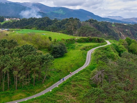Aerial view of a winding road through lush green hills and dense forests with people walking near the Silence Beach Viewpoint in Cudillero Asturias Spain.