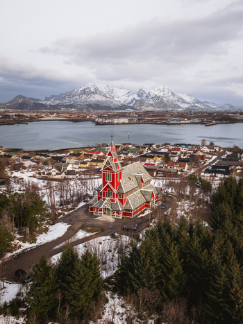 Aerial view of the striking red Buksnes Church stands boldly against the muted winter landscape framed by snow dusted trees and distant mountains Buksnes Kirke Nordland Norway. Aerial view of the striking red Buksnes Church stands boldly against the muted winter landscape framed by snow dusted trees and distant mountains Buksnes Kirke Nordland Norway.
