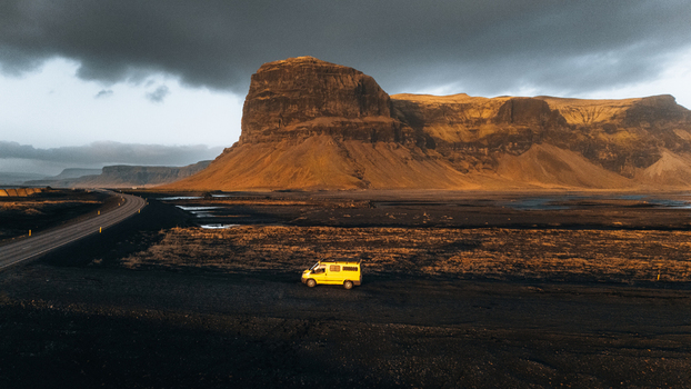 Aerial view of the Lomagnupur mountain with a yellow van parked on the dark volcanic plains under a dramatic sky in Skaftarhreppur Iceland.