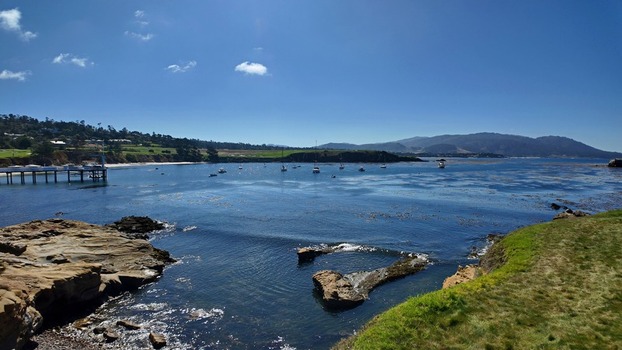 View of 17 Mile Drive coastline with boats anchored in the bay a wooden pier rocky foreground and green hills under a clear blue sky in Del Monte Forest California United States. View of 17 Mile Drive coastline with boats anchored in the bay a wooden pier rocky foreground and green hills under a clear blue sky in Del Monte Forest California United States.