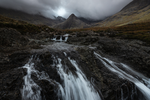View of Fairy Pools with cascading waterfalls over dark rocks under a dramatic cloudy sky and mountain peaks Isle of Skye Scotland United Kingdom.