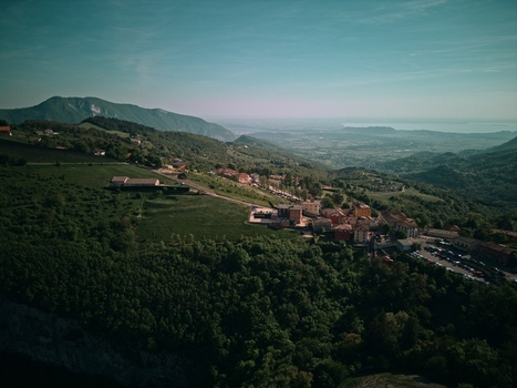 Aerial view of Lake Garda in the distance with a small hillside village surrounded by dense green forests and rolling mountains under a clear sky Italy.