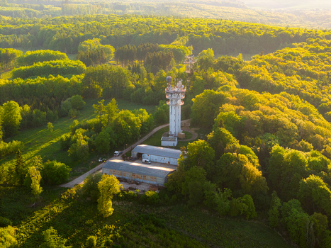 Aerial view of a white telecommunications tower surrounded by dense green forest and small utility buildings during golden hour in Chvalkovice Hradec Kralove Region Czechia.