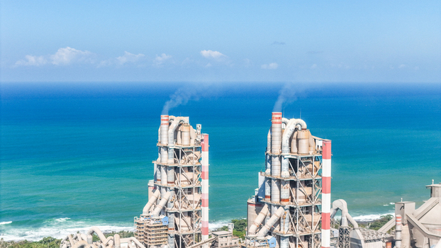 Aerial view of a large industrial cement plant with two tall towers emitting smoke situated along the coastline of Indonesia.