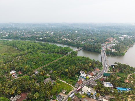 Aerial view of the Gin Ganga river bridge surrounded by lush tropical greenery palm trees and residential houses under a hazy sky in Galle Southern Province Sri Lanka.