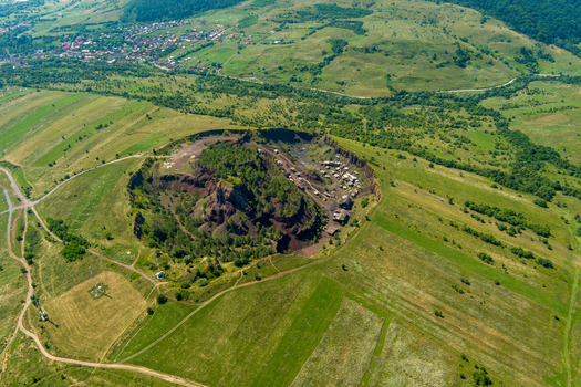 Brasov Romania 21 September 2026 Aerial view of the Racos volcanic crater with its unique red and black volcanic rock formations surrounded by vibrant green meadows and rolling hills. Brasov Romania 21 September 2026 Aerial view of the Racos volcanic crater with its unique red and black volcanic rock formations surrounded by vibrant green meadows and rolling hills.