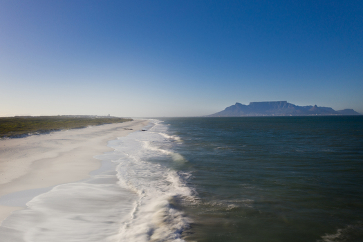 Aerial view of the long white sandy coastline with crashing waves and Table Mountain on the horizon under a clear blue sky in Cape Town Western Cape South Africa. Aerial view of the long white sandy coastline with crashing waves and Table Mountain on the horizon under a clear blue sky in Cape Town Western Cape South Africa.