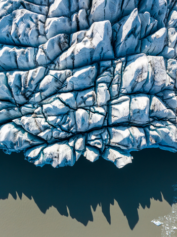 Aerial view of a massive glacier with deep crevasses and textured ice formations casting shadows over the dark glacial water in Iceland. Aerial view of a massive glacier with deep crevasses and textured ice formations casting shadows over the dark glacial water in Iceland.