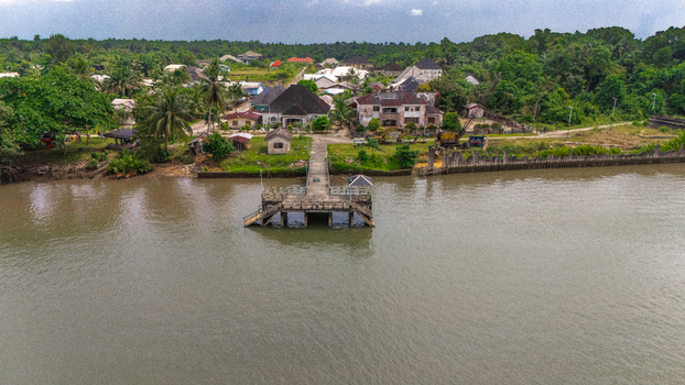 Aerial view of a concrete pier extending into a river with houses lush green trees and vegetation along the riverbank in Rivers Nigeria.