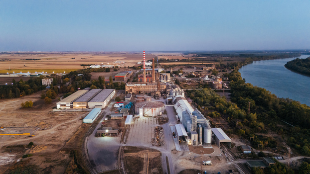 Aerial view of an industrial factory complex situated along the riverbank featuring large storage silos chimneys and surrounding agricultural fields in Sremska Mitrovica Vojvodina Serbia. Aerial view of an industrial factory complex situated along the riverbank featuring large storage silos chimneys and surrounding agricultural fields in Sremska Mitrovica Vojvodina Serbia.