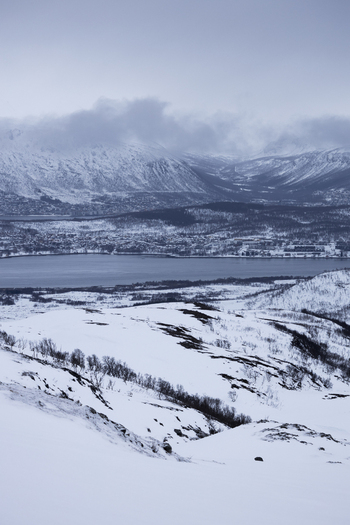 View of snow covered mountains and a coastal settlement along the fjord under a misty winter sky Kvaloya Troms Norway.