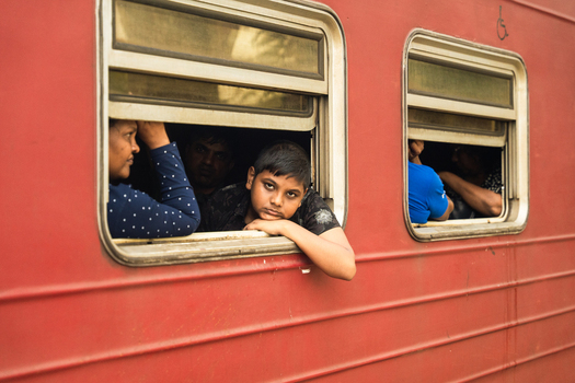 Sri Lanka 06 February 2026 View of passengers looking out from the windows of a red train carriage featuring a young boy leaning on the window sill.