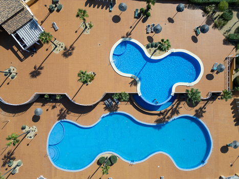 Aerial view of two uniquely shaped blue swimming pools surrounded by a tiled patio palm trees and sun umbrellas in Moraira Valencian Community Spain.