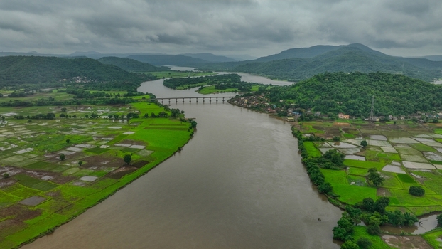 Aerial view of the Mumbai Goa Highway bridge spanning a wide river surrounded by vibrant green agricultural fields and rolling hills in Mahad Maharashtra India.