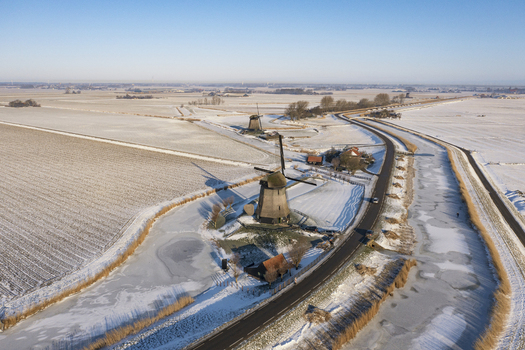 Aerial view of a traditional Dutch windmill standing in a snow covered landscape beside a frozen canal and a road in Ursem North Holland Netherlands.