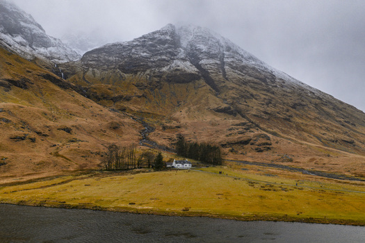 Aerial view of the white cottage and snow dusted mountains in the rugged landscape of Glencoe Scotland United Kingdom. Aerial view of the white cottage and snow dusted mountains in the rugged landscape of Glencoe Scotland United Kingdom.