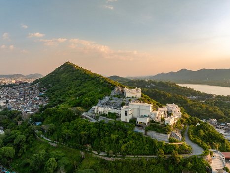 Aerial view of the hilltop palace surrounded by lush green hills rocky terrain and a nearby lake under a warm sunset sky in Udaipur Rajasthan India.