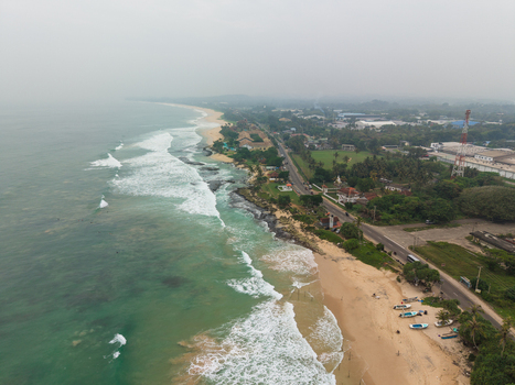 Aerial view of the sandy beach with crashing waves coastal road and lush tropical vegetation under a misty sky Koggala Southern Province Sri Lanka. Aerial view of the sandy beach with crashing waves coastal road and lush tropical vegetation under a misty sky Koggala Southern Province Sri Lanka.