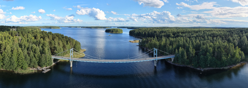 Aerial view of Potkusalmen silta Bridge spanning the calm waters of a lake surrounded by dense pine forests under a blue sky with scattered clouds in Savonlinna Etela Savo Finland.