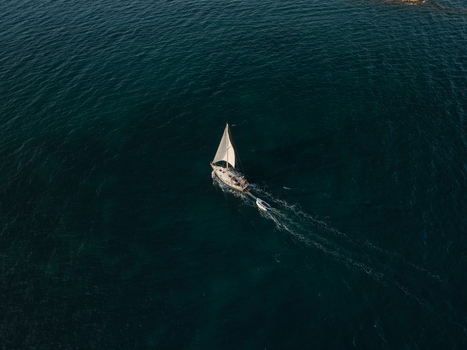 Aerial view of a sailboat with white sails and a small boat in tow cruising on the deep blue sea Syracuse Sicily Italy. Aerial view of a sailboat with white sails and a small boat in tow cruising on the deep blue sea Syracuse Sicily Italy.