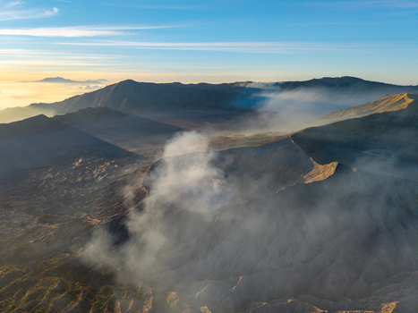 Aerial view of Mount Bromo at sunrise with volcanic smoke rising from the crater rugged terrain and golden light illuminating the surrounding mountains in Tosari East Java Indonesia. Aerial view of Mount Bromo at sunrise with volcanic smoke rising from the crater rugged terrain and golden light illuminating the surrounding mountains in Tosari East Java Indonesia.