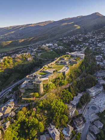 Aerial view of Gjirokaster Fortress perched on a hill overlooking the historic stone city with lush greenery and mountains in Gjirokaster Gjirokaster County Albania. Aerial view of Gjirokaster Fortress perched on a hill overlooking the historic stone city with lush greenery and mountains in Gjirokaster Gjirokaster County Albania.