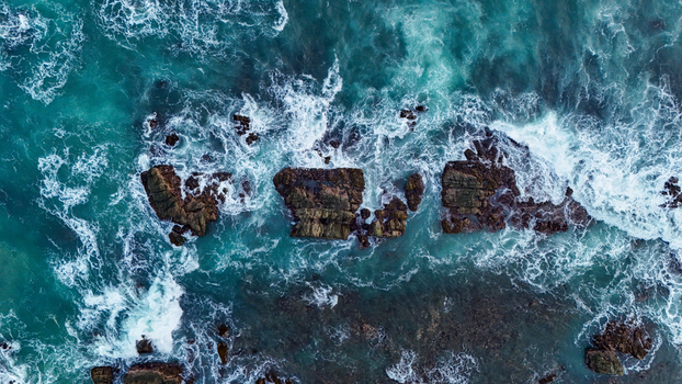 Aerial view of waves crashing against dark rocks along the turquoise coastline of Indonesia. Aerial view of waves crashing against dark rocks along the turquoise coastline of Indonesia.