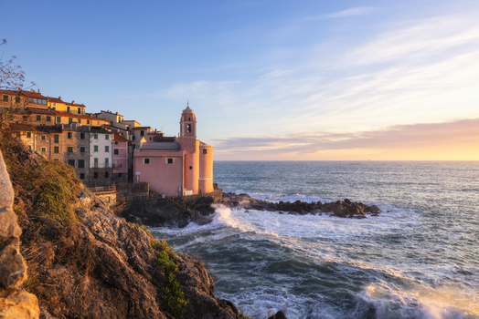 View of the pink Church of San Giorgio on the rocky coastline with waves crashing and colorful houses at sunset in Tellaro Liguria Italy. View of the pink Church of San Giorgio on the rocky coastline with waves crashing and colorful houses at sunset in Tellaro Liguria Italy.