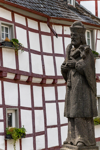 View of a stone statue of a religious figure in front of a traditional half timbered house with white walls and dark wooden beams Monreal Rhineland Palatinate Germany. View of a stone statue of a religious figure in front of a traditional half timbered house with white walls and dark wooden beams Monreal Rhineland Palatinate Germany.