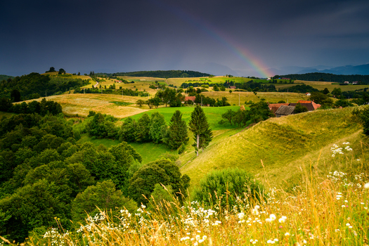 View of rolling green hills and meadows with a farmhouse nestled in the landscape under a dramatic stormy sky with a vibrant rainbow in Brasov Brasov Romania.
