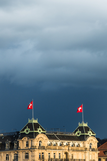 Zurich Switzerland 02 April 2023 View of Swiss flags atop a grand building contrasting sharply against the ominous dark sky above. Zurich Switzerland 02 April 2023 View of Swiss flags atop a grand building contrasting sharply against the ominous dark sky above.