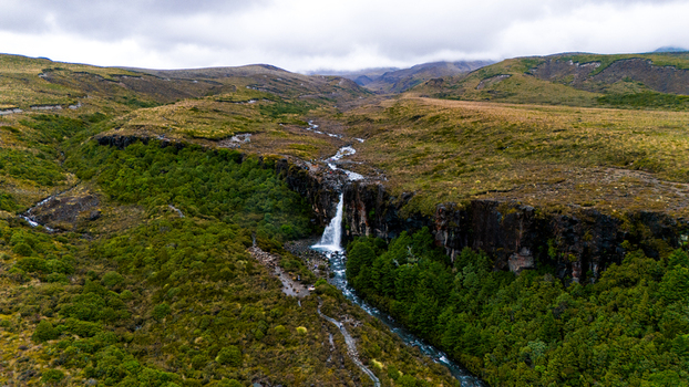 Aerial view of Taranaki Falls cascading down a dark rocky cliff into a verdant gorge against a backdrop of rolling hills under a cloudy sky Taranaki Falls Manawatu Whanganui Region New Zealand. Aerial view of Taranaki Falls cascading down a dark rocky cliff into a verdant gorge against a backdrop of rolling hills under a cloudy sky Taranaki Falls Manawatu Whanganui Region New Zealand.
