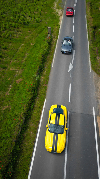 Aerial view of three sports cars including a yellow Mustang with black stripes driving along a rural road through lush green vineyards in La Chapelle de Guinchay Bourgogne Franche Comte France.
