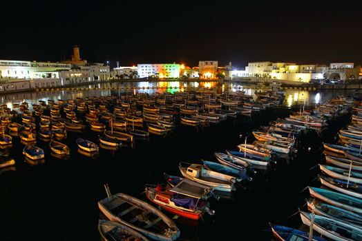 View of numerous small fishing boats moored in a calm harbor at night with illuminated buildings along the waterfront in Algeria.